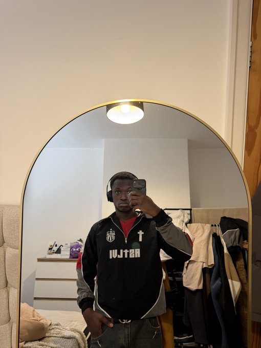A man takes a mirror selfie in a bedroom, wearing headphones and a black athletic jacket with 'IV.I.T.2.H' text and a cross logo.