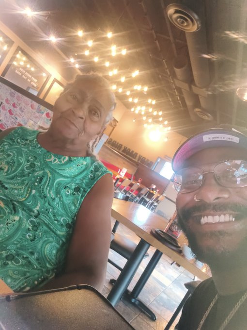 A selfie of a smiling man wearing glasses and a cap with "LBW JERSEY" next to an older woman in a green paisley top, taken inside a brightly lit restaurant.