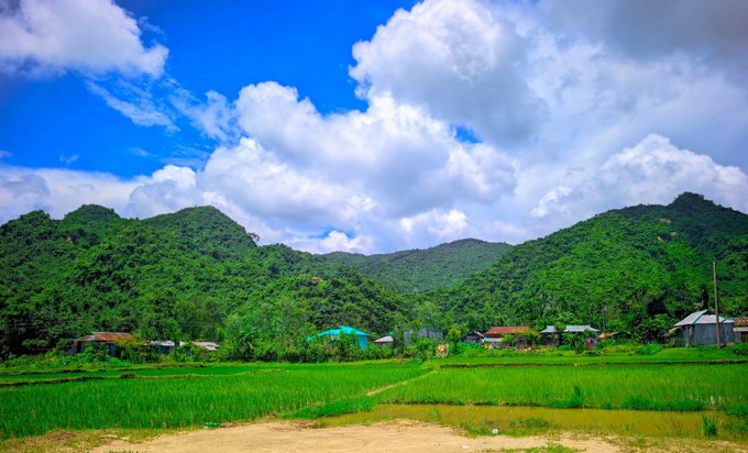 A scenic landscape shows lush green mountains, rice paddies, and a small village under a bright blue sky with puffy white clouds.