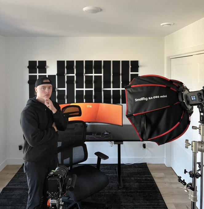 A young man in a black hoodie stands in a home studio setup with a curved monitor, acoustic foam panels, and a SmallRig RA-D60 mini softbox light.