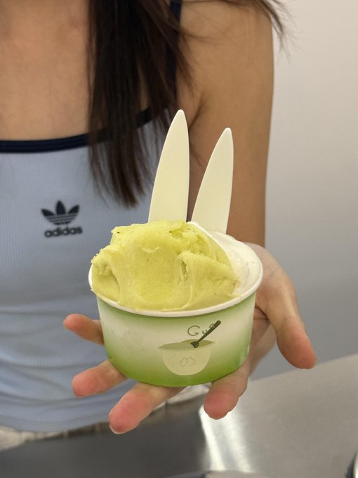 A hand holds a cup of green ice cream/dessert decorated with white ear-shaped pieces, worn by a person in a light blue Adidas crop top.