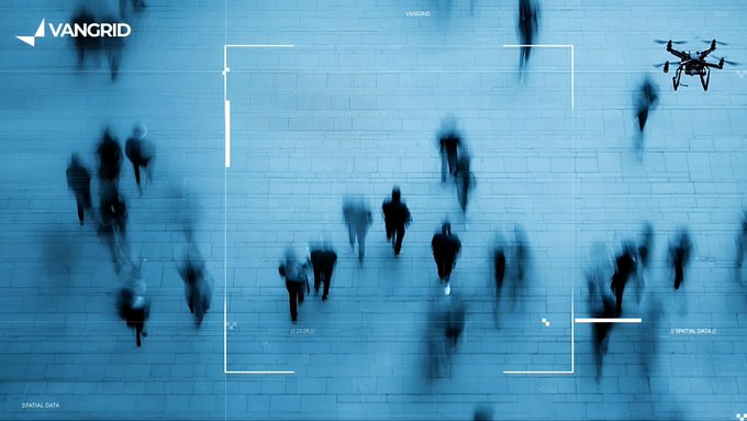 A blue-tinted aerial surveillance view of pedestrians walking on a plaza, framed by a tactical camera overlay with a drone visible in the upper right, branded 'VANGRID'.