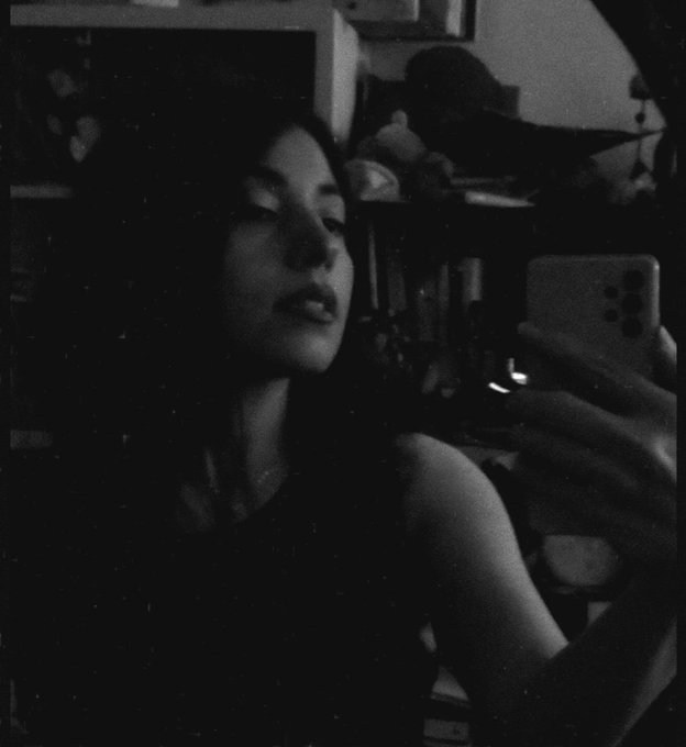 A moody, grainy black-and-white mirror selfie of a young woman with dark hair, holding a smartphone and gazing into a dimly lit room filled with shelves and objects, paired with a morning greeting celebrating the first sale of her NFT artwork "Self-Portrait #7" (edition 2/5, 35 XTZ) on objkt.com. The intimate, film-like aesthetic of the self-portrait complements the personal tone of the post, serving as both a visual representation of the sold artwork and a candid expression of gratitude to the collector.