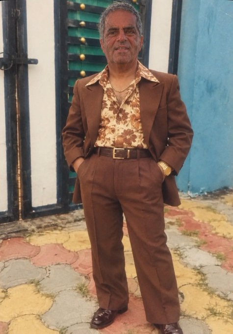 An old photograph of a smiling man in a brown suit and floral shirt posing outside a building with a green door and colorful tiled pavement.