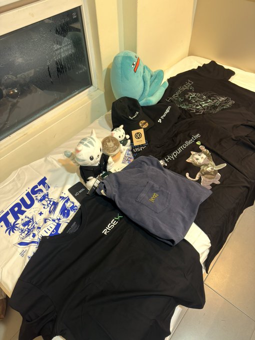 Various merchandise including t-shirts with 'TRUST' and 'RISE' logos, plush toys, and hats are arranged on a bed.