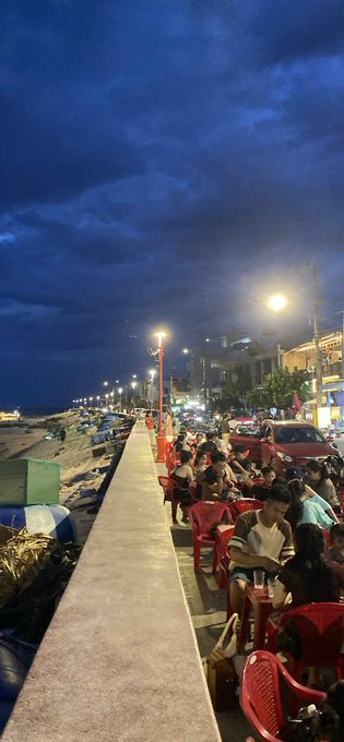 A nighttime scene at a crowded beachfront promenade where people sit at red plastic chairs under streetlights along the seaside.