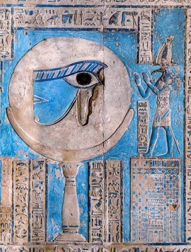 A photograph of an ancient Egyptian stone carving featuring the Eye of Horus set within a disc, painted in blue, with hieroglyphics surrounding it and a deity figure on the right.