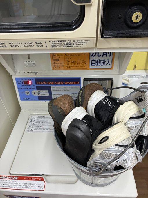 A bucket filled with New Balance sneakers sits inside a Japanese 'COIN SNEAKER WASHER' machine, with Japanese text and pricing visible on the machine.