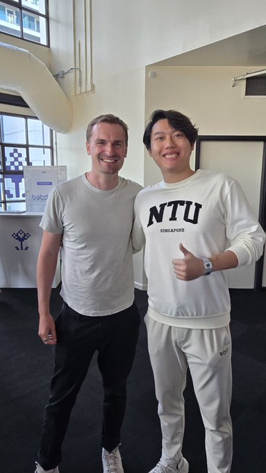 Two men smile and pose together indoors; the man on the right wears a white sweatshirt with 'NTU SINGAPORE' printed on it and gives a thumbs-up.