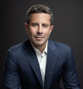 A professional headshot of a man with dark hair wearing a dark navy suit jacket over a white collared shirt, posed against a dark gray background.