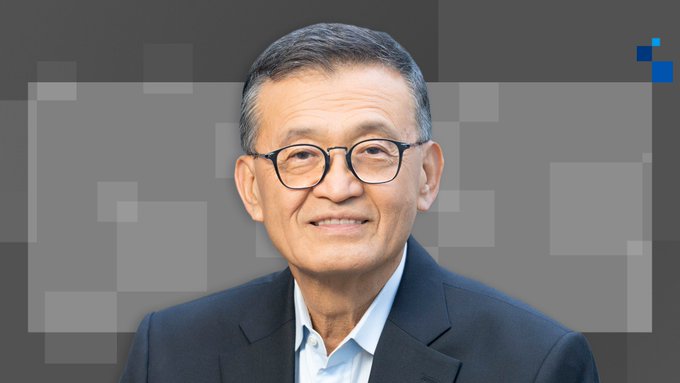 The image features a man wearing glasses, sitting next to a large square. He appears to be smiling at the camera with his hands resting on his lap or behind him. The backdrop of the photo has a pattern resembling a grid layout, giving off an intellectual or formal vibe. In the background, there is text that reads "@intel has started risk production of 18A-P", which may have a connection to the man's name or the content of the image in some way. Additionally, a line of code with letters and numbers appears to be part of the grid layout behind him.