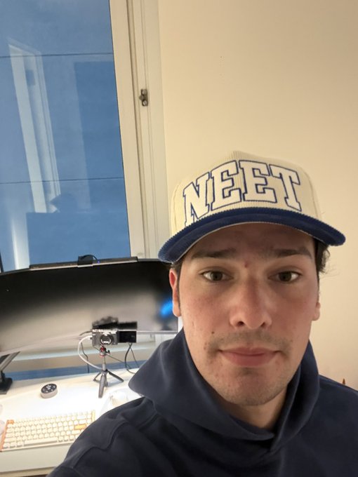 A man wearing a white and blue "NEET" snapback hat and a navy hoodie takes a selfie at his streaming setup, featuring a microphone and keyboard visible in the background. The image ties to his post celebrating NEET culture while promoting his Twitch stream focused on catching the next 8-figure runner in the onchain market.