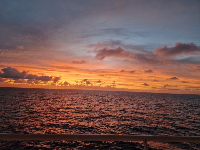 A vibrant sunset over the open ocean, with orange, pink, and purple hues reflecting on the rippling water, viewed from behind a ship's railing.
