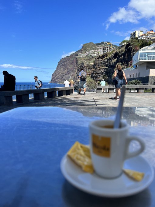 A scenic coastal view featuring a cup of espresso and a waffle cookie on a saucer in the foreground, with tourists walking along a promenade beside the ocean and rocky cliffs.