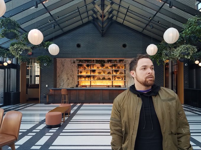A bearded man in a black hoodie and olive green bomber jacket stands in a stylish hotel lobby with a checkered floor, hanging globe lights, and a warmly lit bar in the background.