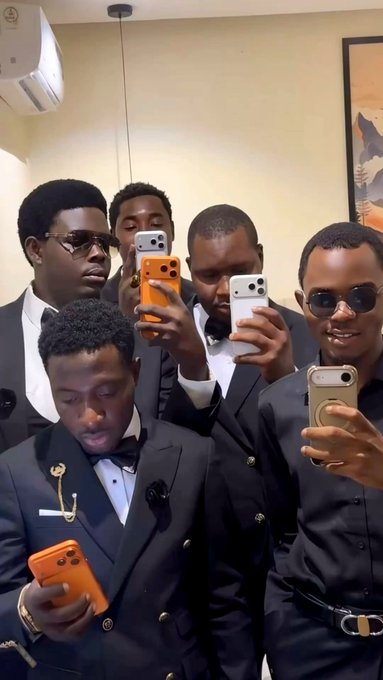 A group of five men in formal black suits pose together, with several holding up smartphones to take a mirror selfie.