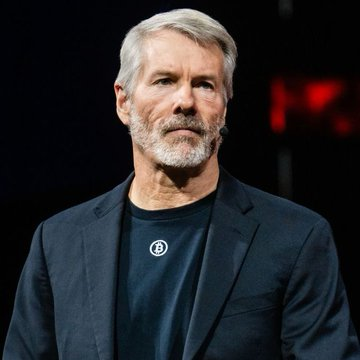 Michael Saylor, executive chairman of MicroStrategy ($MSTR), is shown in a dark blazer over a black t-shirt, representing the company whose stock purchase by President Trump is being announced in the breaking news post.
