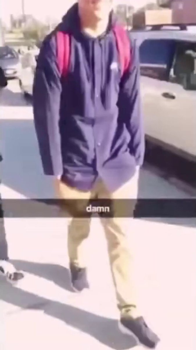 The image shows a man walking down a sidewalk, wearing a blue hoodie with a backpack over his shoulders. The photo has been edited to include text that reads "DAMN" at the bottom of the frame. This suggests that someone might have altered or added this text to the original image for some reason.