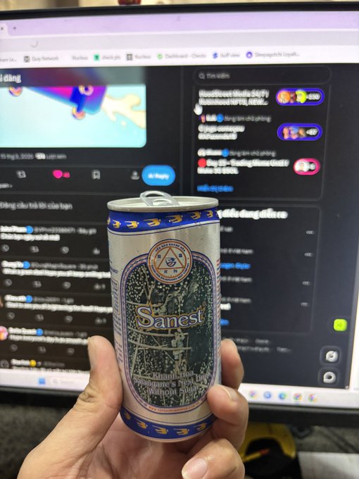 A person's hand holding a blue and silver Sanest beer can in front of a computer monitor displaying what appears to be a social media feed or chat interface with a video playing.