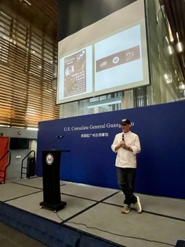 A man giving a presentation at a podium in front of a blue backdrop that reads 'U.S. Consulate General Guangzhou' with a large projection screen displaying slides behind him.