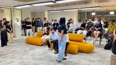 An indoor event space where a group of attendees sits and stands around yellow modular seating, with people appearing to engage in discussion or a presentation.