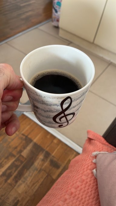 A hand holding a white mug with sheet music design and a treble clef, filled with black coffee, over a wooden floor with an orange blanket visible.