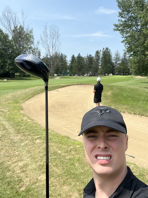 A man wearing a Toronto Blue Jays cap grimaces in the foreground while a golf club stands upright in a sand trap; another person in black walks in the distance on a sunny golf course.