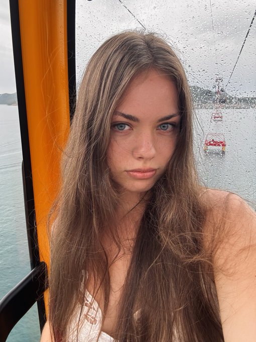A young woman with long brown hair takes a selfie inside a cable car, with raindrops on the window and another gondola visible in the misty background, fitting her casual "gm" morning greeting to her followers.