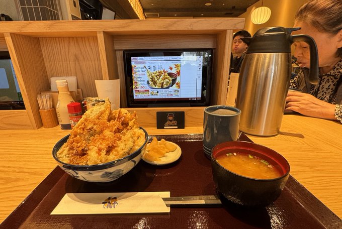 A meal at a Japanese restaurant featuring a bowl of rice topped with fried items, miso soup, and a thermos, with a tablet menu displaying prices like '740円' and '800円' in the background.