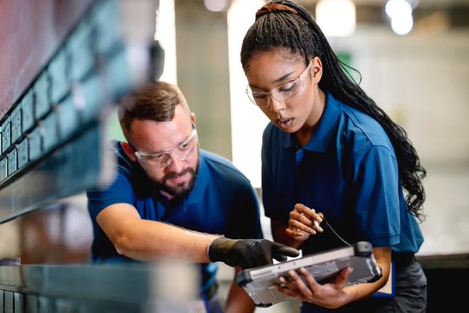 A photograph of a male and a female engineer in a manufacturing facility, both wearing blue work shirts and safety glasses, examining a laptop computer.