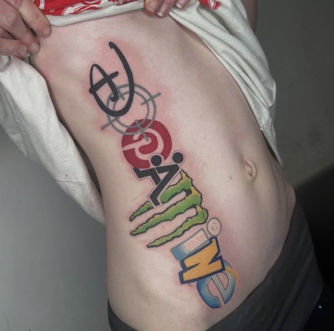 A person's torso with a colorful vertical tattoo featuring overlapping symbols including a circle with a cross, a crosshair target, a red shape, a '7', a green Christmas tree, and the number '5'.