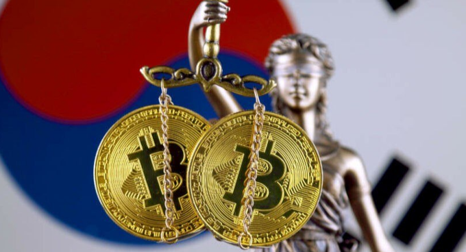 Two Bitcoin coins displayed as pendants in front of a Lady Justice statue with the South Korean flag in the background.