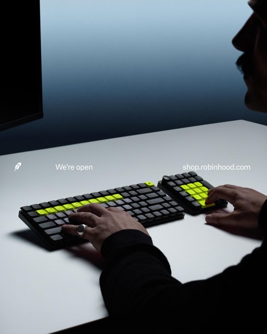 A man types on a split ergonomic keyboard with neon-yellow highlighted keys in a dimly lit setting, with the text "We're open" and "shop.robinhood.com" displayed on a white desk. The accompanying post playfully suggests the man resembles Vlad Tenev, CEO of Robinhood, and notes an apparent "A3" tattoo on his finger, hinting at deliberate symbolism related to the company's branding and market activity.