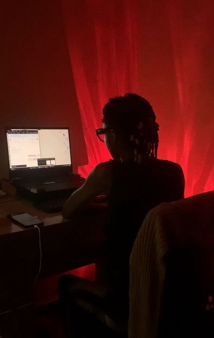 A person with glasses sits at a desk using a laptop in a dimly lit room bathed in red light from a curtain behind them.