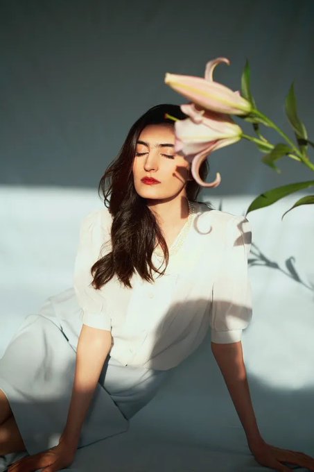 Woman in white clothing sitting in sunlight with her eyes closed, with a pink lily flower positioned near her face.