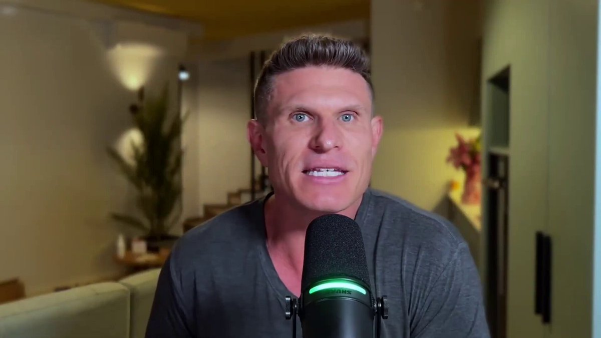 The image shows a man wearing a gray shirt sitting behind a microphone, likely preparing for a live stream or recording session. The background features a green wall with a potted plant placed near it. There are also two chairs visible in the scene, one on each side of the man and the microphone.