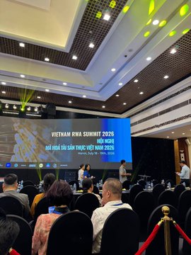 A conference room showing the 'VIETNAM RWA SUMMIT 2026' event with attendees seated facing a presentation screen displaying 'MỞ HỚI TÀI SẢN THỰC, VIỆT NAM 2026'.