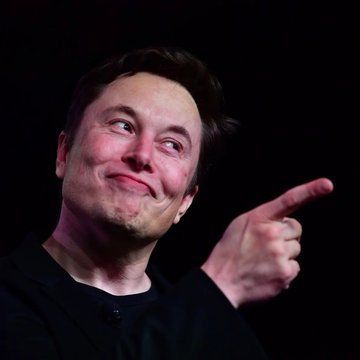 The image features a man with brown hair wearing a black shirt who is pointing his finger towards something out of frame. The background appears to be dark, which contrasts with the bright light shining on the man's face as he makes eye contact with the camera. There are no other people or objects in the image besides the man and the point of focus.
