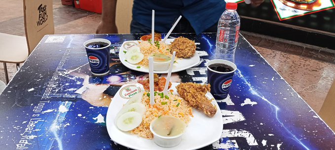 The image shows two plates of food sitting on a table, along with cups of soda. The table also has a poster or advertisement for a Chinese restaurant that includes a picture of fried chicken. A person can be seen standing near the table, possibly waiting to enjoy their meal.