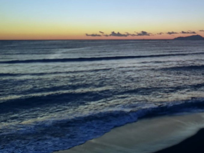 Ocean waves roll onto a sandy shore at sunset, with distant mountains visible under a soft pink and orange sky. The serene seascape captures the tranquil moment of "waiting for waves," mirroring the anticipation of the "Waves" NFT collection launch.