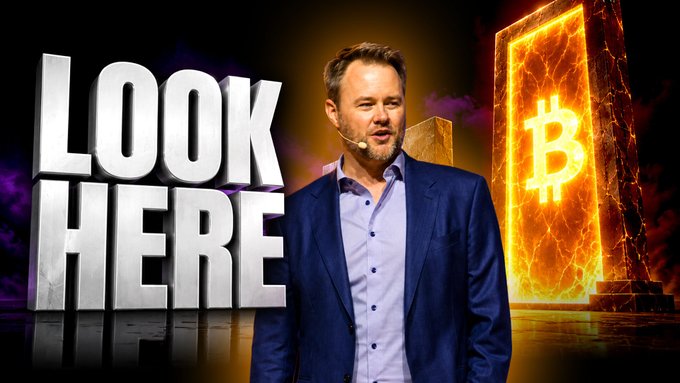 A man in a blue suit stands with a microphone in front of large 3D text reading 'LOOK HERE' and a glowing Bitcoin logo on a fiery background.