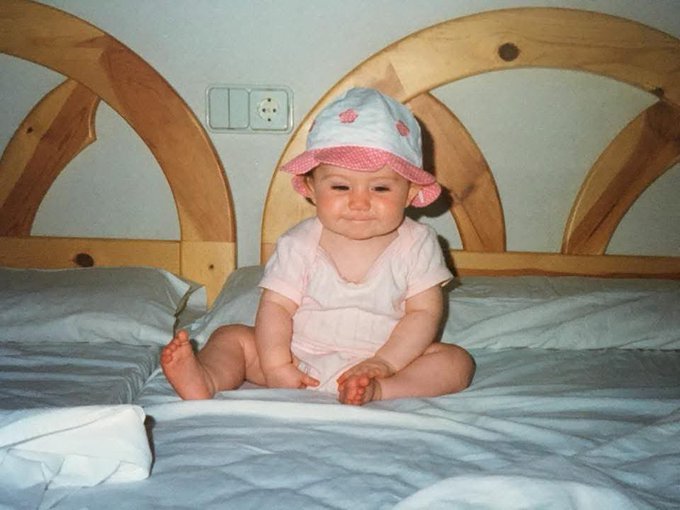 A vintage photograph of a smiling baby wearing a pink sun hat with polka dots and a pink outfit, sitting on a bed with a wooden arched headboard.