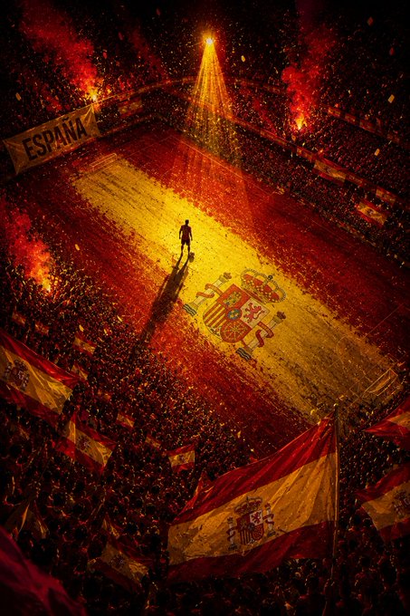 An aerial view of a massive stadium crowd displaying a giant Spanish flag with the national coat of arms, illuminated by a central spotlight on a lone figure, with smaller Spanish flags and an 'ESPAÑA' banner visible.