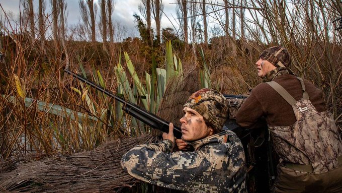 Two hunters in camouflage attire lie in wait in a marsh, their shotguns ready, mirroring the anticipation and stillness mentioned in the post about Bitcoin's current market behavior.