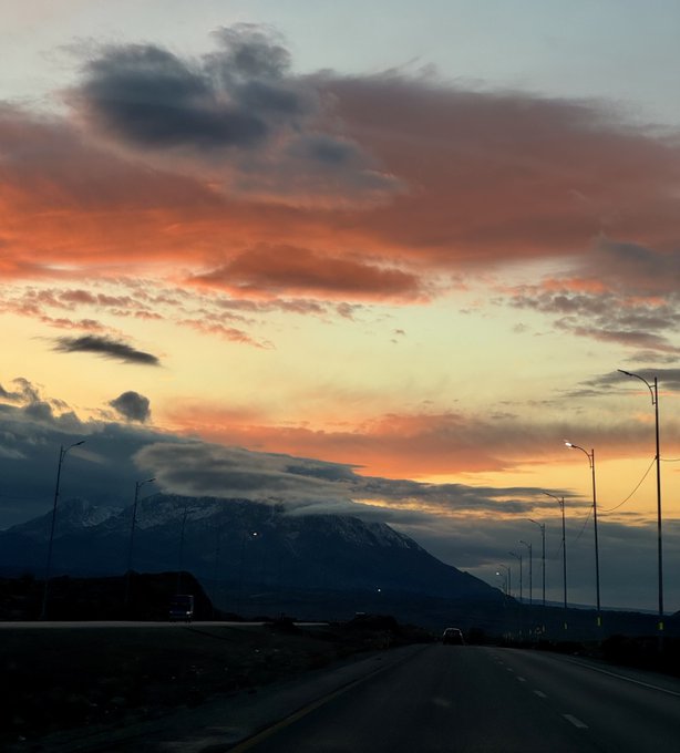 A scenic view from a highway at sunset, with orange and pink clouds filling the sky above snow-capped mountains.