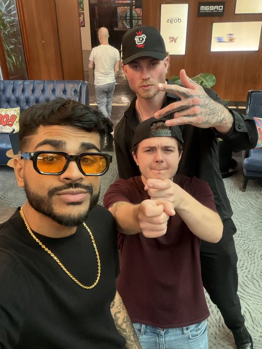 Three young men pose for a selfie indoors; one wears yellow-tinted sunglasses and a gold chain, while another wears a black cap with a red logo.