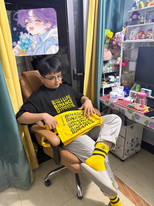 A young man wearing glasses, a black Binance t-shirt, and a yellow Binance bag, sitting in an office chair in a room decorated with anime figures, plush toys, and an anime poster.