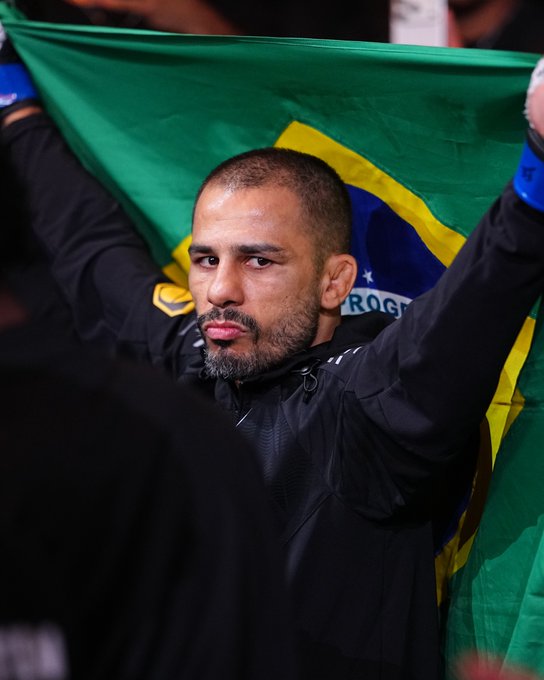 A bearded fighter holds up the Brazilian flag, with "ROGR" visible on it, capturing a moment of pride and resilience that matches the caption's tribute to Pantoja's warrior spirit at UFC 331.