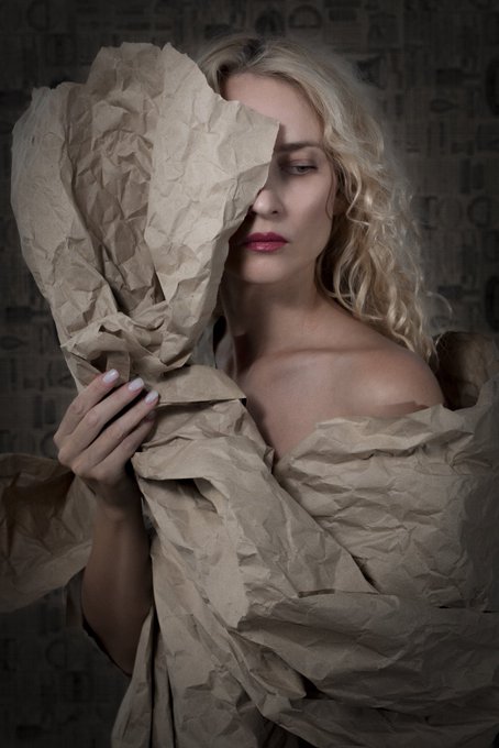 A portrait of a blonde woman with bold red lipstick, her face partially obscured by crumpled brown craft paper she holds up against a dark, textured background.
