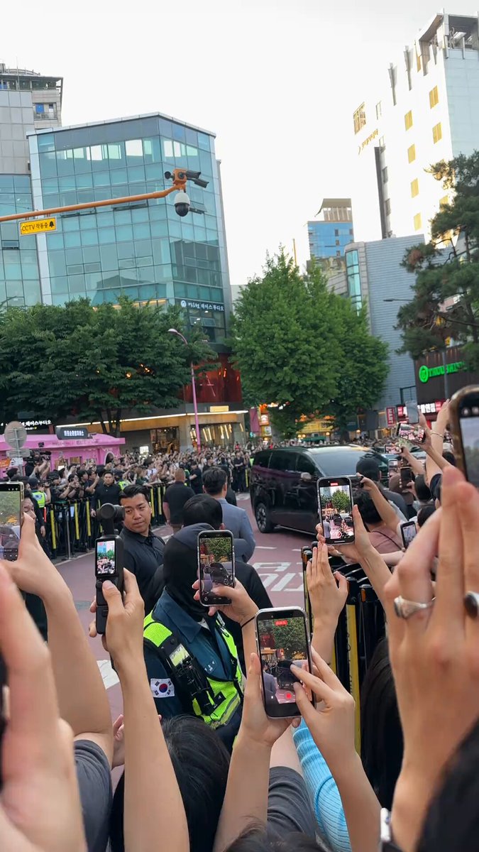 The image shows a large crowd of people gathered around a man who appears to be rushing towards them, with security personnel surrounding him. The crowd consists of at least thirteen individuals holding their cell phones up high, capturing the moment as they watch the man approaching. The scene suggests that there might have been an incident or an event that has drawn the attention of the people in the area.